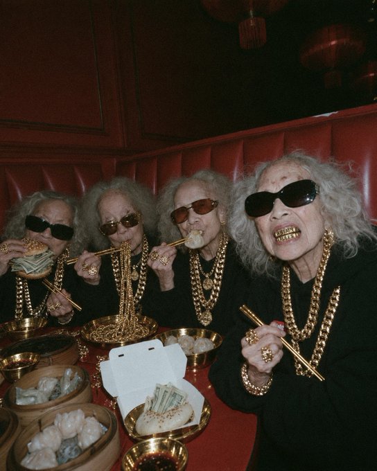 A stylized photo of four older women wearing sunglasses and heavy gold chains and jewelry, eating dumplings and drinking from gold-rimmed cups at a restaurant booth.