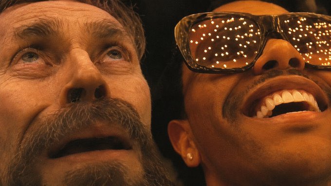 A close-up of two men looking upward; the man on the right wears sunglasses reflecting numerous small lights.