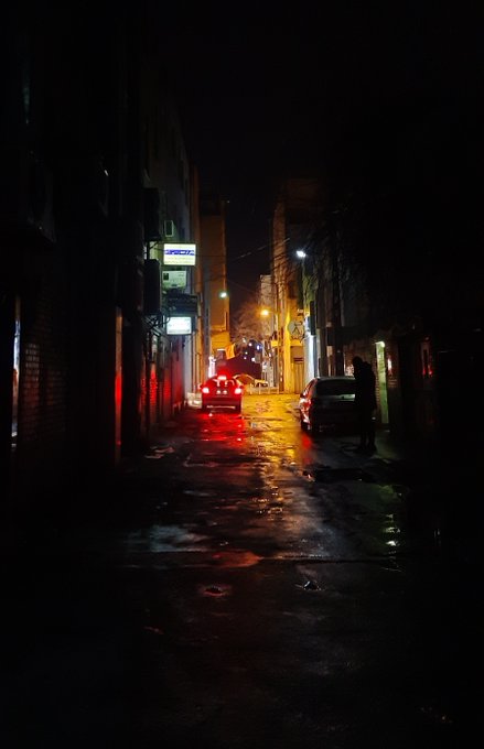 A dark, atmospheric night scene of a narrow alley or street illuminated by the red glow of car taillights, with a solitary figure standing near a parked vehicle.