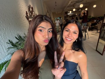 Two women pose for a selfie, smiling and flashing peace signs in what appears to be a restaurant or cafe setting.