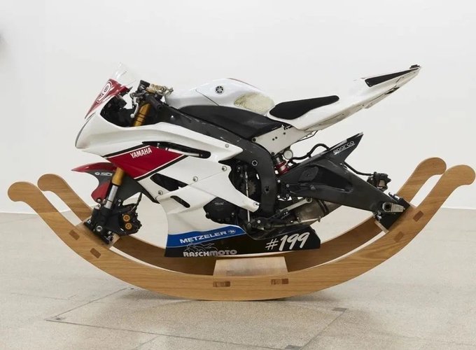 A white and red Yamaha racing motorcycle is creatively mounted on a wooden rocking chair base, displayed in a gallery setting with the number '#199' visible on its side.