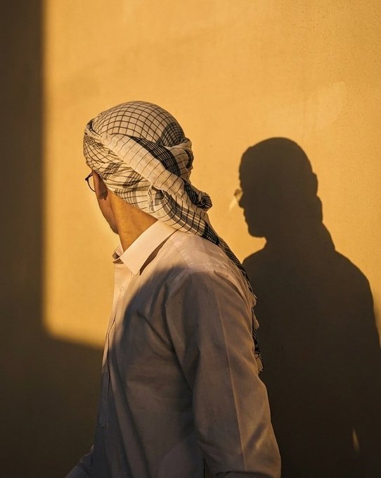 A man in a checkered keffiyeh and white shirt stands in profile against a warm, sunlit wall, casting a sharp silhouette of his head and shoulders. The post's mention of "Habibti dress" connects to the elegant, traditional Middle Eastern attire featured in the image.