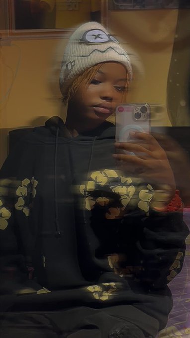The image features a young woman wearing a black hoodie with yellow flowers on it, taking a selfie using her cell phone. The background includes a mirror that reflects the girl's face as well as other people present in the room. There are also two instances of the word "Mutual" visible in the image.