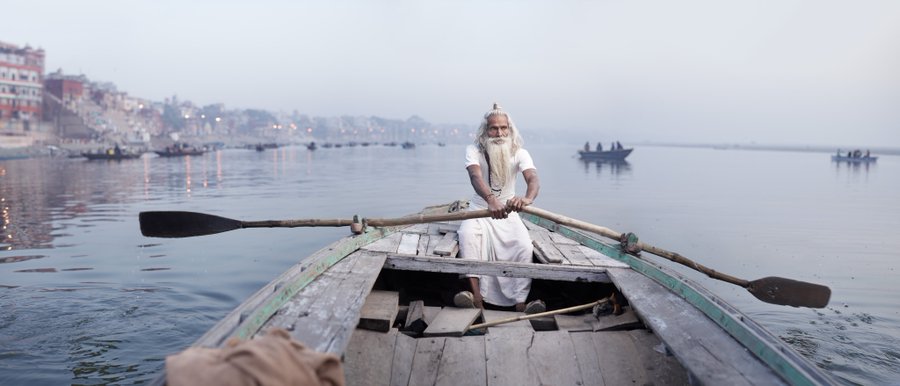 An elderly man with a long white beard rows a wooden boat on a misty river, with ghats and other boats visible in the background.