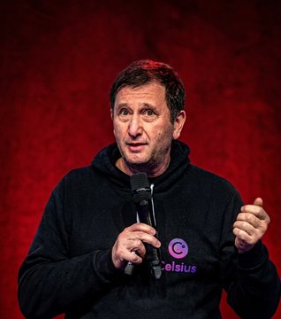 The image features a man standing in front of a red background, holding a microphone and speaking into it. The man appears to be wearing a black sweatshirt with a purple logo on it. There are also words visible in the image that read "CFTC permanently bans former Celsius CEO Alex Mashinsky from its regulated markets". This suggests that the man is addressing an audience or participating in a public event, possibly related to cryptocurrency regulation and the ban of certain individuals within the industry.