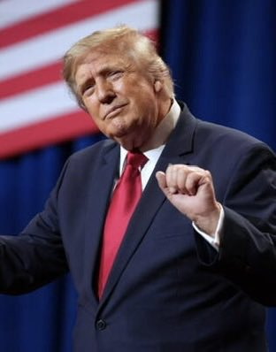 Donald Trump gestures with a clenched fist in front of an American flag backdrop, wearing a dark suit and red tie. The image accompanies news about his cryptocurrency ($TRUMP) surging 2,700% while the parody $LAPTOP coin (referencing the Biden laptop controversy) dropped 98%, highlighting the dramatic divergence between the two politically-themed meme coins shortly after launch.
