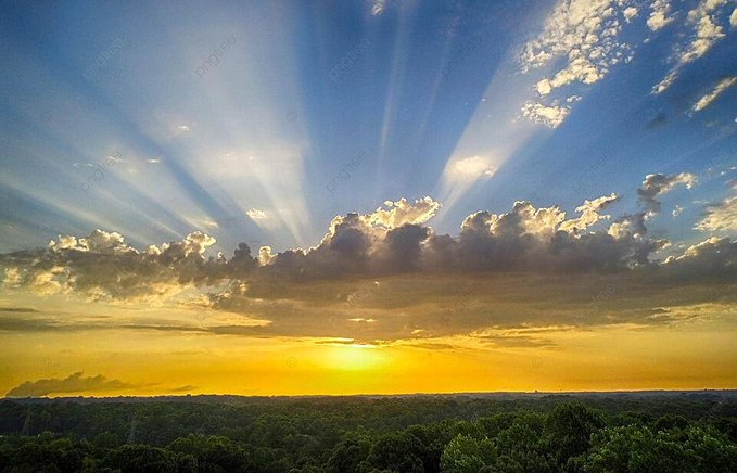 The image shows a beautiful sunset over a forested area with trees, clouds, and sunlight shining through them. The sun appears to be setting behind the trees, creating a warm glow across the scene. The sky is filled with bright rays of light from the sun, casting long shadows on the ground below. This picturesque view captures the essence of nature's beauty and tranquility.
