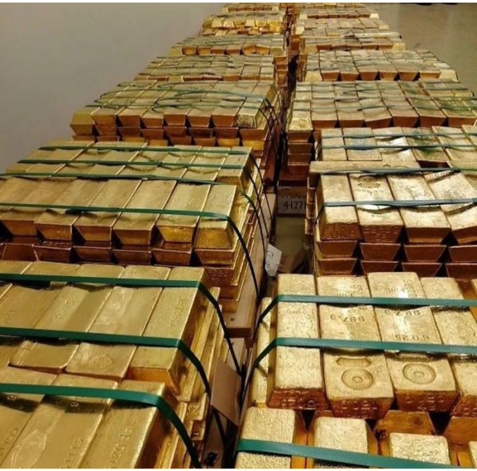Rows of stacked, shiny gold bullion bars are tightly bound with green plastic straps, arranged in a warehouse setting. The bars display various stamped numbers and markings on their surfaces.