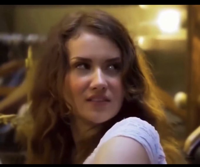 A close-up shot of a young woman with curly brown hair smiling subtly, looking off-camera in what appears to be a warmly lit indoor setting.