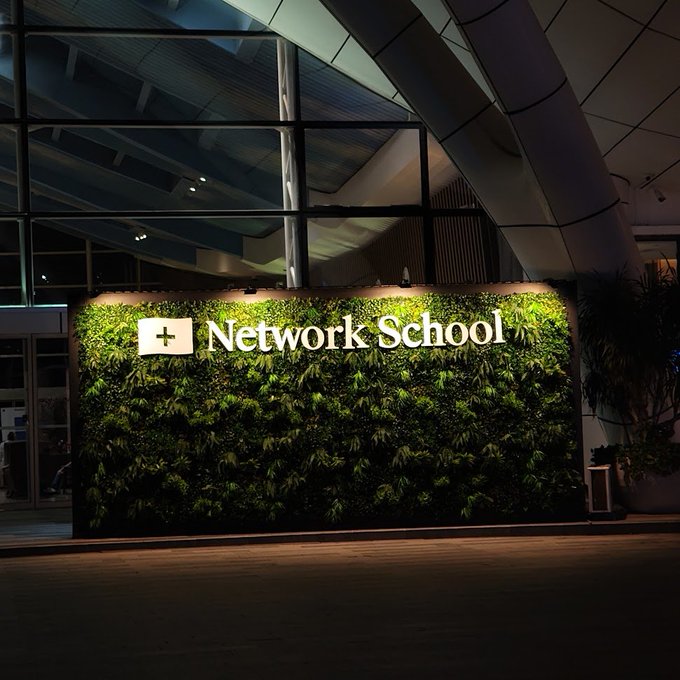 The image shows a large green wall with the words "Network School" written on it, likely located inside an office building or a school. The wall has many plants growing on it, adding a touch of nature to the indoor space. The photo is taken at night, highlighting the unique design and lighting conditions within the building.