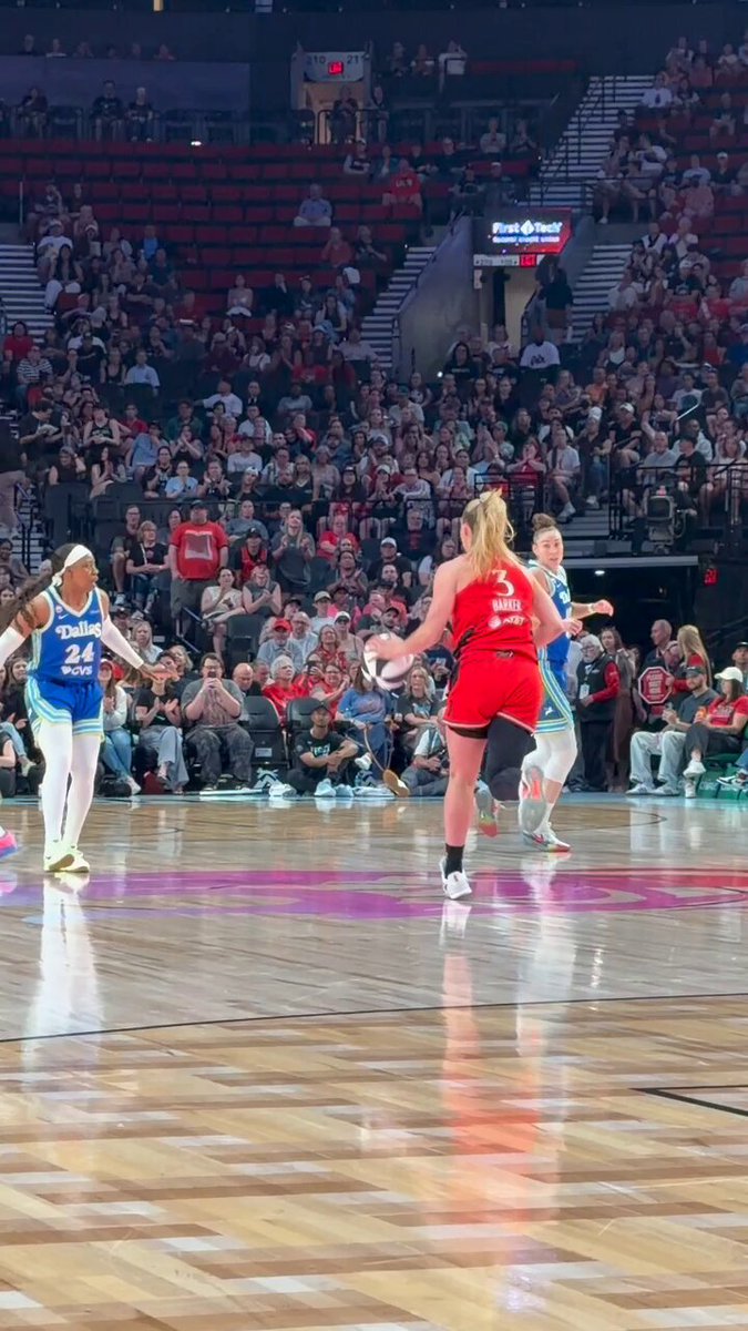 The image shows a basketball court with two women playing against each other, one wearing a red shirt and the other wearing blue. The court has a wooden floor that reflects the light from above. In the background, there are several people watching the game, some of whom appear to be sitting in chairs while others stand around the court. A sign is visible on the wall behind them, which reads "@coinbase Tap to watch: wnba.com/how-to-watch-g".