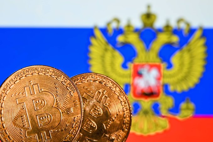 Two gold Bitcoin tokens rest in the foreground against a blurred backdrop of the Russian flag and the Russian Federation's double-headed eagle coat of arms. Visible engravings on the coins include "BITCOIN DIGITAL," "DECENTRALIZED," and "PEER TO PEER," visually reinforcing the article's announcement that Russia's central bank is proposing to allow Bitcoin, Ethereum, and USDT to be publicly traded on official exchanges starting September 1, 2026.