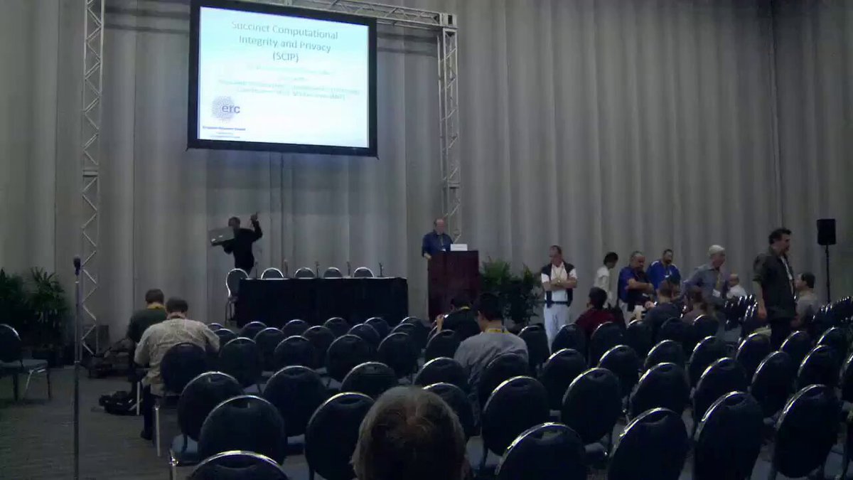The image shows an indoor event with a large screen displaying information about cryptocurrency. A man stands behind a podium at the front of the room, likely presenting or discussing the topic. The audience consists of people seated in rows of chairs facing the stage. There are potted plants placed around the room, adding to the ambiance and decorating the space.