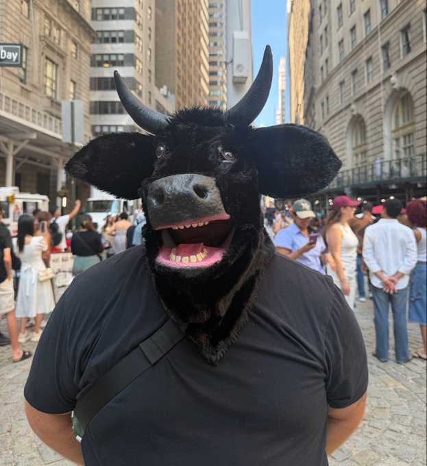 A person wearing a black bull mask with horns and a wide, toothy grin stands in a crowded city street, with a partial "dev" sign visible on a building in the background. The post's caption humorously suggests the masked "bull" is celebrating winning 5000 $ANSEM, tying the bullish imagery to the meme coin win.
