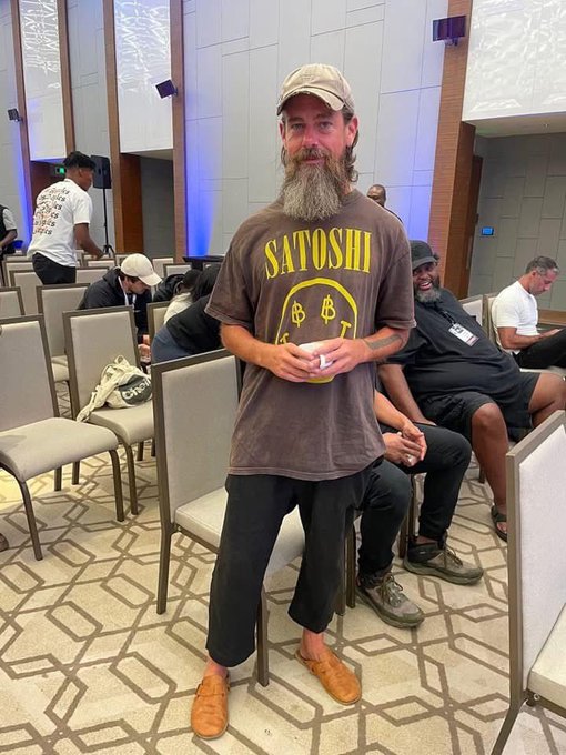 A bearded man wearing a tan cap and a brown 'SATOSHI' Bitcoin t-shirt stands in a conference room holding a cup, with other attendees seated behind him.
