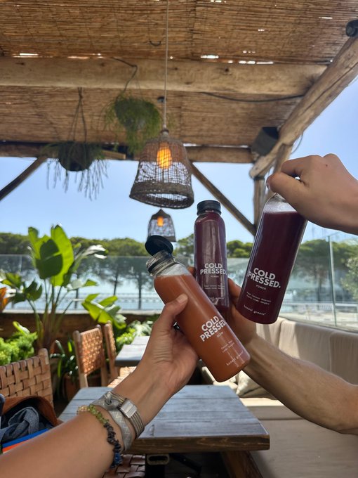 Three hands clink bottles and a glass of 'Cold Pressed' beverages together in a cheers gesture at an outdoor patio with a beach view.