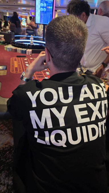 A man sits at a casino gaming table wearing a black t-shirt with the white text 'YOU ARE MY EXIT LIQUIDITY' printed on the back.
