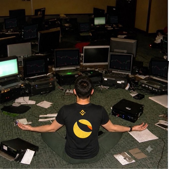 A man in a t-shirt featuring the Binance "B" logo sits in a meditative lotus pose, surrounded by an array of trading monitors displaying financial charts and scattered paperwork. His calm, centered posture amid the chaotic trading setup visually reinforces the post's mantra "Hold and breathe," embodying the patience required during volatile market activity.