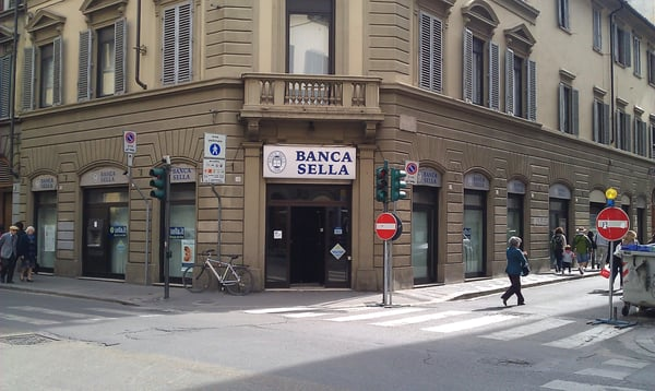 The image shows a street corner with a building that has "Banca Sella" written on its facade, indicating that this is an Italian bank. The bank recently became the first Italian bank approved under MiCA to offer crypto custody services, which means they are now part of the European banking system and can provide services related to cryptocurrency management.

There are several people walking around in front of the building, including a person crossing the street at a crosswalk. A bicycle is parked nearby, suggesting that someone might have used it for transportation or exercise. The presence of these elements indicates an active urban environment where people go about their daily activities.