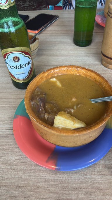 A bowl of soup with meat and cheese chunks served in a rustic clay bowl on a red and blue plate, accompanied by a bottle of 'Presidente' beer and a green glass on a wooden table.