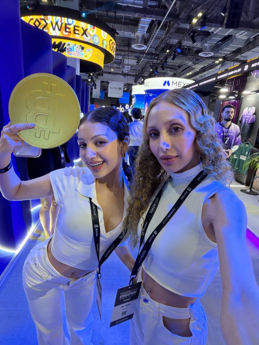 Two women pose at the Token2049 crypto conference, one holding a large gold Bitcoin coin, with "WEEX" and "MEXC" branding visible in the background—capturing the networking and promotional energy that makes the event the year's top crypto gathering.