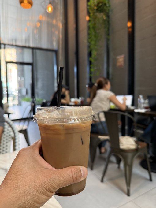 A first-person perspective photo of a hand holding an iced coffee drink with a black straw inside a cafe, with blurred patrons visible in the background.