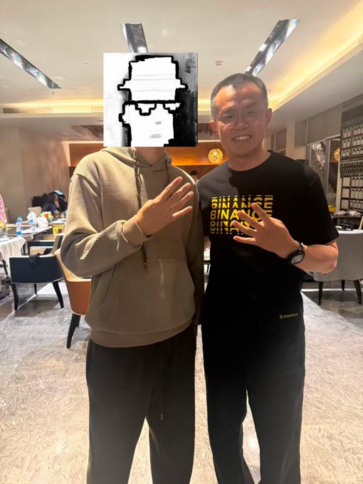 Two men pose for a photo indoors, one with a pixelated face wearing a tan hoodie, and the other wearing glasses and a black 'BINANCE' t-shirt, both making peace signs.