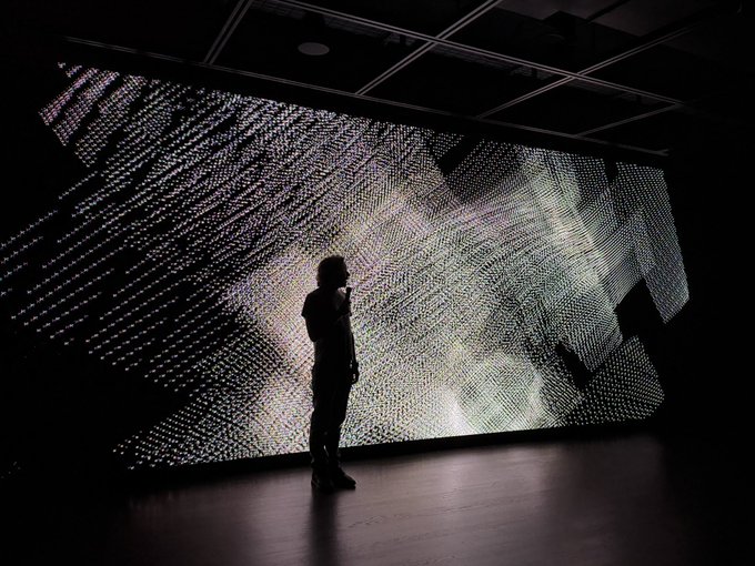 A silhouetted figure stands in a dark room before a large illuminated wall display showing a complex, dotted, wave-like pattern.