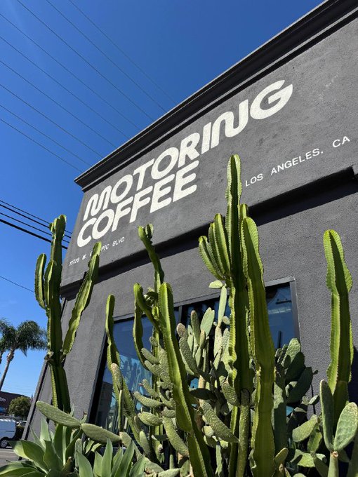 A black building displays "MOTORING COFFEE" in large white letters alongside "LOS ANGELES, CA," with lush cacti in the foreground, capturing the venue for the Cars, Crypto & Coffee event.