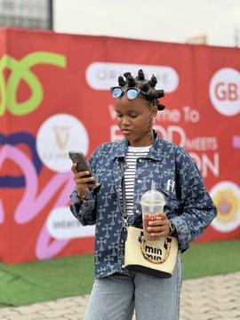 A young woman with hair curlers and sunglasses on her head stands outside a red backdrop, looking at her phone while holding a drink and a 'mini donuts' bag.