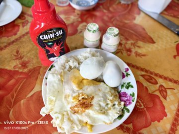 A breakfast plate containing fried eggs and toast sits on a floral tablecloth next to a bottle of Chin-Su chili sauce and two small spice jars, taken with a vivo X100s Pro.