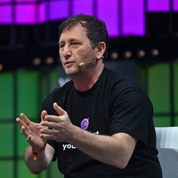 Alex Mashinsky, co-founder of Celsius, is captured mid-speech wearing a black t-shirt with a "C" logo and a headset microphone, gesturing expressively during what appears to be a panel discussion. The image accompanies news of his permanent ban from the securities, commodities, and crypto industries by NY Attorney General Letitia James, along with a $35M financial penalty.