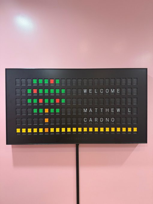 A black electronic LED panel mounted on a pink wall displaying the message 'WELCOME MATTHEW L CARDNO' alongside a colorful pixelated pattern, branded 'MASTABOARD' at the bottom.