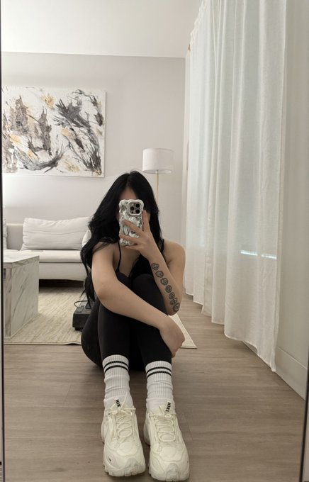 A mirror selfie of a person with long dark hair sitting on a wooden floor in a black outfit and chunky white sneakers, wearing white striped crew socks, with tattoo-like markings visible on their arm.