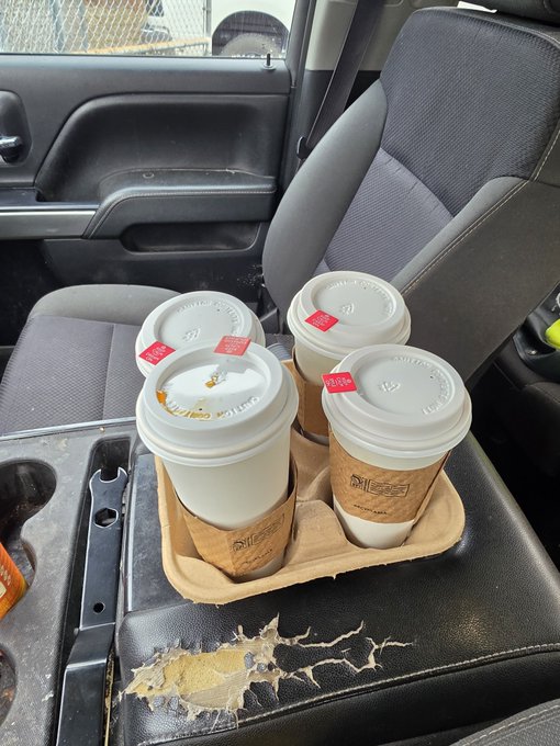 Three Starbucks coffee cups with red 'ORDER HERE' stickers sit in a cardboard tray on the center console of a car, where the surrounding black leather is visibly worn and peeling.