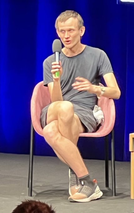 A man in a gray t-shirt and shorts sitting in a pink chair on stage, holding a microphone, resembling Vitalik Buterin.