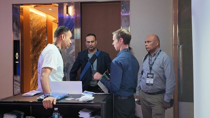 Four men are gathered around a counter in what appears to be a conference or hotel setting, with one man in a white shirt leaning on a counter with a laptop.