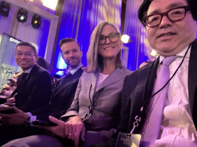 The image shows a group of people sitting together, with two men wearing suits and glasses and two women wearing business attire. They are all smiling for a photo, likely at an event or gathering. The background features a stage with blue curtains, suggesting that this could be a formal event or a performance. The caption reads "CATHIE WOOD SOLD A LOT. But not Bitmine. ARK dumped positions across 4 funds right before the SpaceX IPO. $ACHR $BIDU $HOOD 
ARXR XRX $TER $KTOS $IRDM $LHX $XE Not one share of 
BMNR $ETH @fundstrat". The image is a collage of different elements, including people sitting together and the stage with blue curtains.
