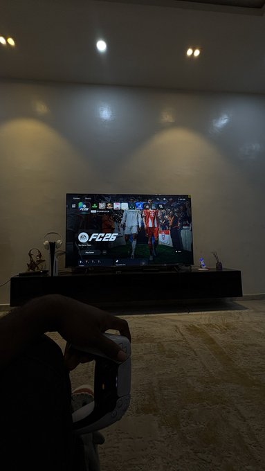 A hand holds a PlayStation controller while a TV displays the EA Sports FC 25 main menu screen, set in a dimly lit living room. The "GM CT" caption likely refers to a casual gaming moment, capturing the relaxed experience of launching the latest football title.