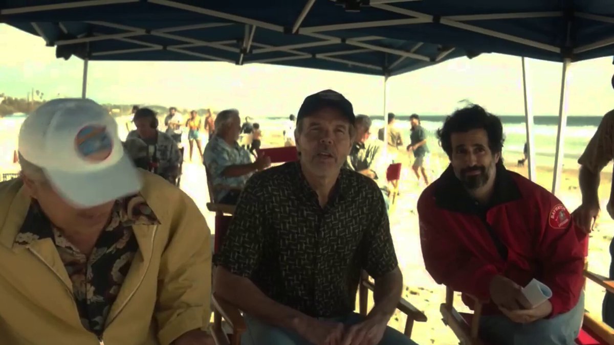 The image shows a group of people sitting under an umbrella, possibly at a beach or a similar outdoor location. There are three men prominently featured in the scene - one wearing a white hat, another wearing a black shirt, and the third wearing a red jacket. The background reveals that they are likely enjoying their time outdoors on a sunny day.