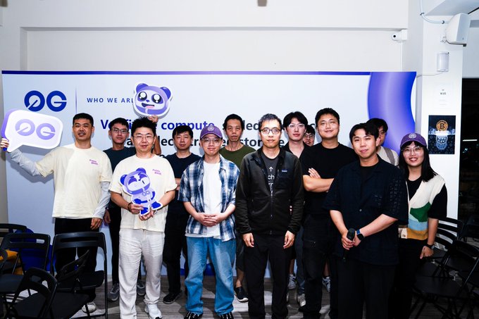 A group photo of about ten people posing in front of a backdrop with 'OG' logos and 'WHO WE ARE' text, with one person holding a sign showing the 'OG' logo.