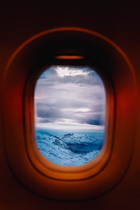 A view through an oval airplane window showing snow-capped mountains, a body of water, and dramatic clouds.