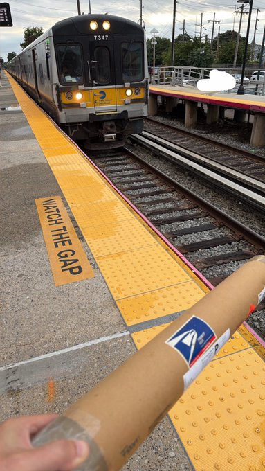 A first-person view at a train station, with a hand holding a cardboard tube in the foreground and a commuter train approaching on the tracks, next to a yellow strip reading "WATCH THE GAP".