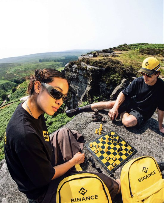 An outdoor photo of two people wearing black Binance-branded clothing and yellow Binance backpacks playing chess on a rocky cliff with a scenic green landscape in the background.