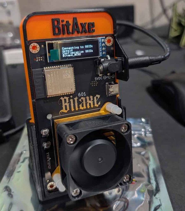 A 'BitAxe 601' cryptocurrency mining device with a small display reading 'Connecting to SSID...', featuring an exposed circuit board, ESP32 chip, and a cooling fan.
