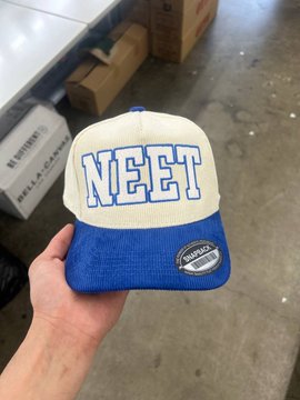 A hand holds a blue and cream corduroy snapback hat featuring the bold "NEET" logo on the front, with a "Snapback" tag on the brim, showcased in what appears to be a warehouse or retail setting—matching the post's excitement about scoring this $NEET branded merchandise.
