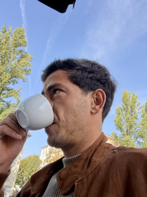 A low-angle selfie of a man with short dark hair and stubble, wearing a brown suede jacket, drinking from a white cup outdoors with trees and a blue sky in the background.
