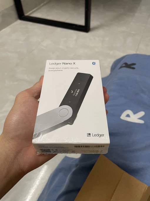 A hand holding a sealed Ledger Nano X hardware wallet box in a room with tiled flooring and what appears to be a shipping package nearby.