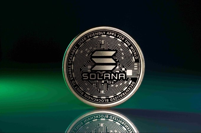 Close-up photograph of a silver Solana (SOL) cryptocurrency coin with 'ENABLE SCALABLE, USER-FRIENDLY APPS FOR THE WORLD' engraved around its edge, sitting on a reflective surface with green lighting.