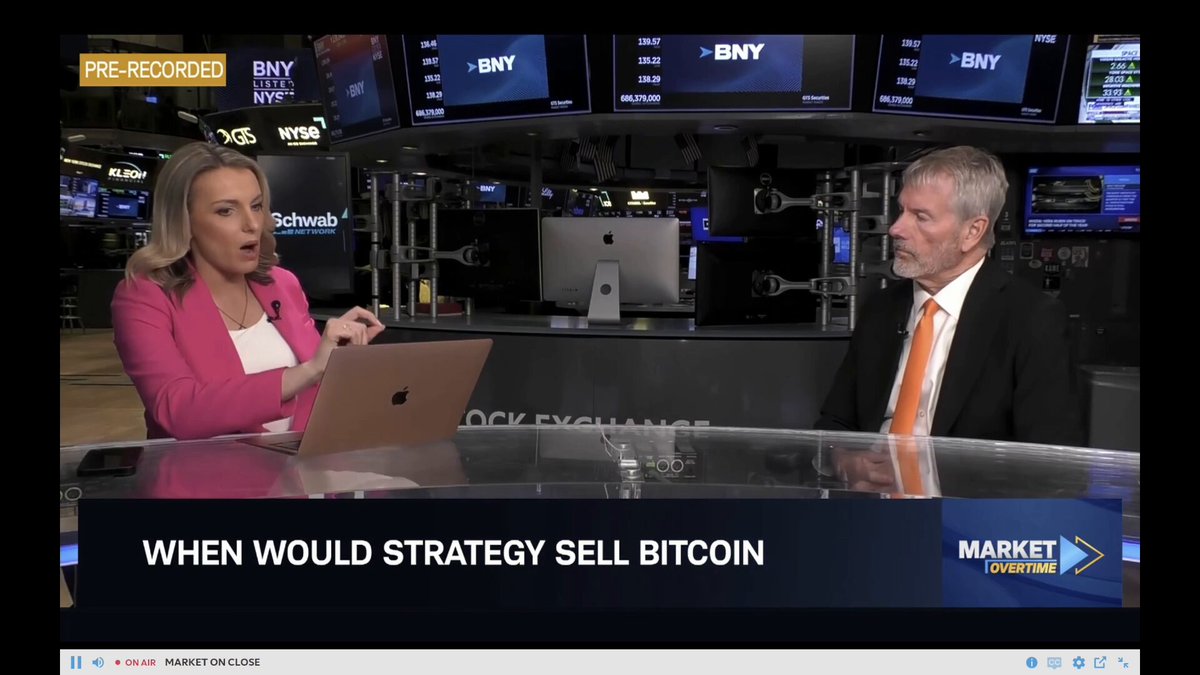The image shows two men dressed in suits seated across from each other behind a desk, engaged in a conversation or interview. The man on the left appears to be a woman, wearing a pink top, while the man on the right is wearing a suit with an orange tie. In front of them, there's a laptop placed on the table and another television screen displaying news about cryptocurrency markets. The post that accompanies this image reads: "MarketWatch: Strategy'S Michael Saylor just said, 'We're at $24 billion run rate with $STRC '. It'is alone going to buy 2-3x Bitcoins produced by miners." The focus of the image is on how these men are discussing strategy and investing in cryptocurrency markets.
